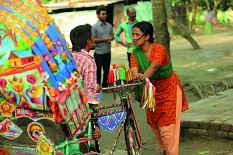 Im Vordergrund ist eine Rikscha zu sehen. Dahinter eine Frau, die mit einem Mann spricht. Beide halten je eine Seite des Fahrradlenkers fest. © Landfilm