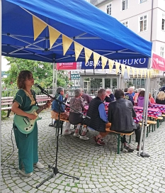 Im Vordergrund steht eine Frau an einem Mikrofon. Sie hält eine Gitarre. Im Hintergrund sind Sitzbänke an Tischen aufgestellt. Auf den Bänken sitzen Menschen. © Rebecca Druschel, i. A. d. Stadt Marburg