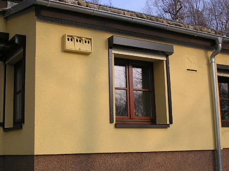 Mattgelbe Hausfassade mit einem Fledermauskasten und einer Sperlingsnisthilfe (beide in der ebenfalls mattgelb)
